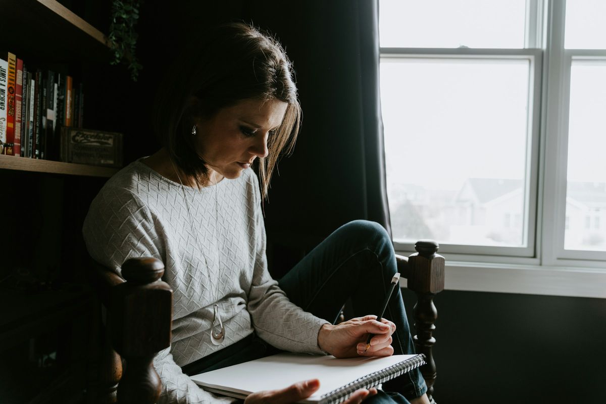A woman sits by a window writing in a notebook.